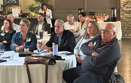 AEM member company representatives gather around a table, listening to a presentation at the most recent AEM Momentum Member Event. 