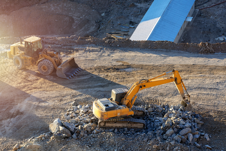 Excavator breaking and moving large rocks at a construction site, with a wheel loader nearby on a graded dirt surface next to a bridge or overpass structure.