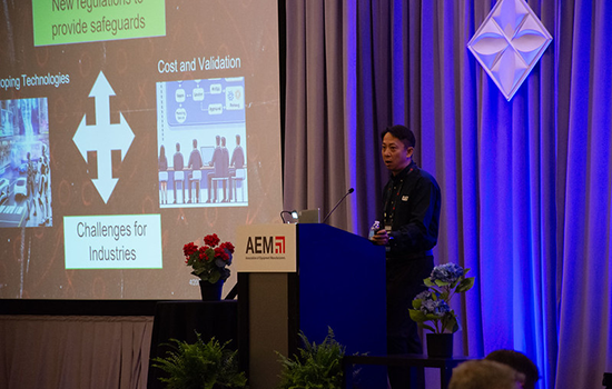 Presenter speaking at an AEM-branded podium beside a slide on industry challenges and cost validation, on a blue-lit stage with curtains, plants, and audience visible in the foreground.