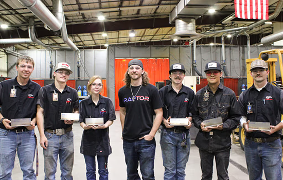 Group of students standing in a training facility, holding their work and posing for a photo in front of industrial equipment and an American flag.