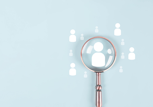 A brass magnifying glass hovers over a group of white profile icons, against a blue background
