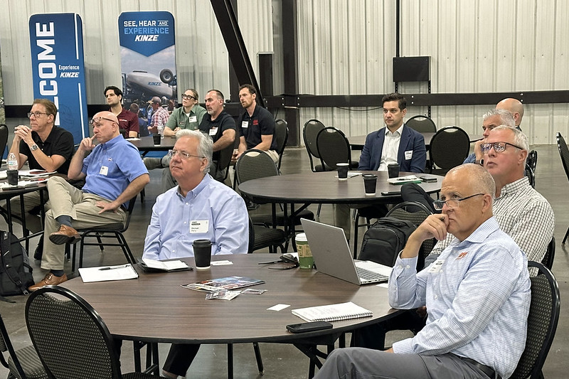 Attendees seated at round tables during a meeting or presentation inside a manufacturing facility, with Kinze-branded banners visible in the background.