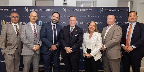 Group of seven business professionals standing in front of a C.D. Howe Institute backdrop, dressed in formal attire and posing for a group photo.