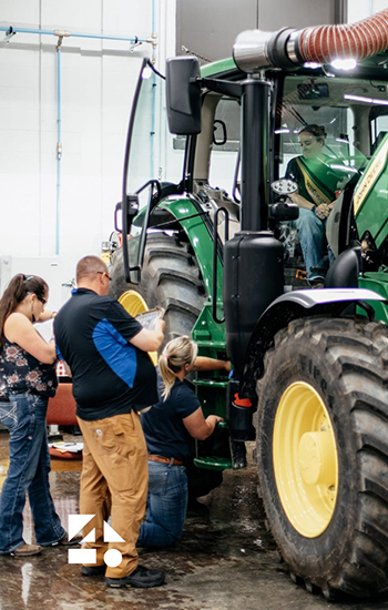 People working together around a large green tractor in an indoor workshop, with one person seated inside the cab while others examine and work on the lower side near the wheel.