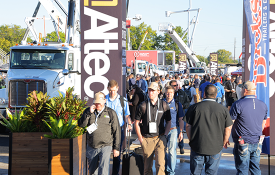 Crowded outdoor Utility Expo show walkway lined with large equipment displays and banners, with attendees walking between booths.