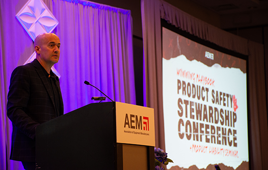 Person speaking at a podium with an “AEM” sign during a conference, with a large screen in the background displaying “Product Safety & Stewardship Conference” and event branding under purple lighting.