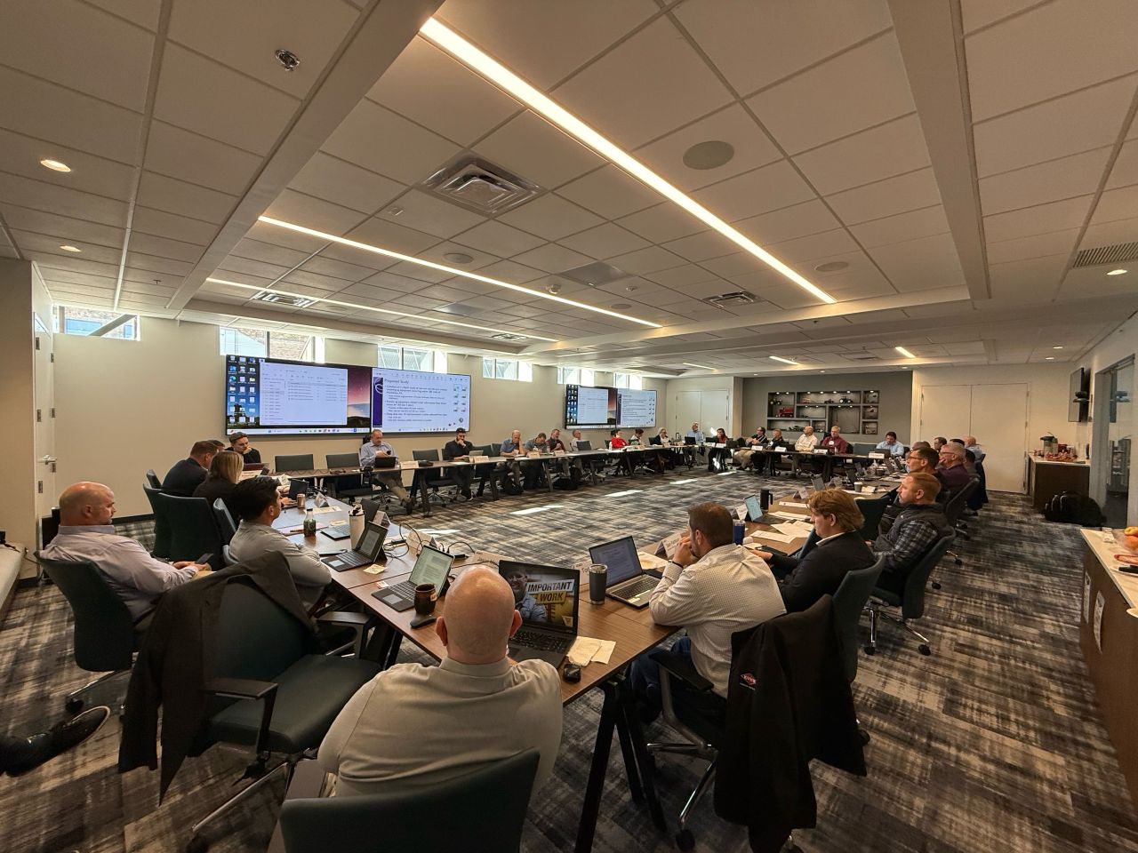 Attendees seated around U-shaped conference tables during AEM's Safety & Product Leadership Council Week in a large conference room, with presentation screens displayed at the front.